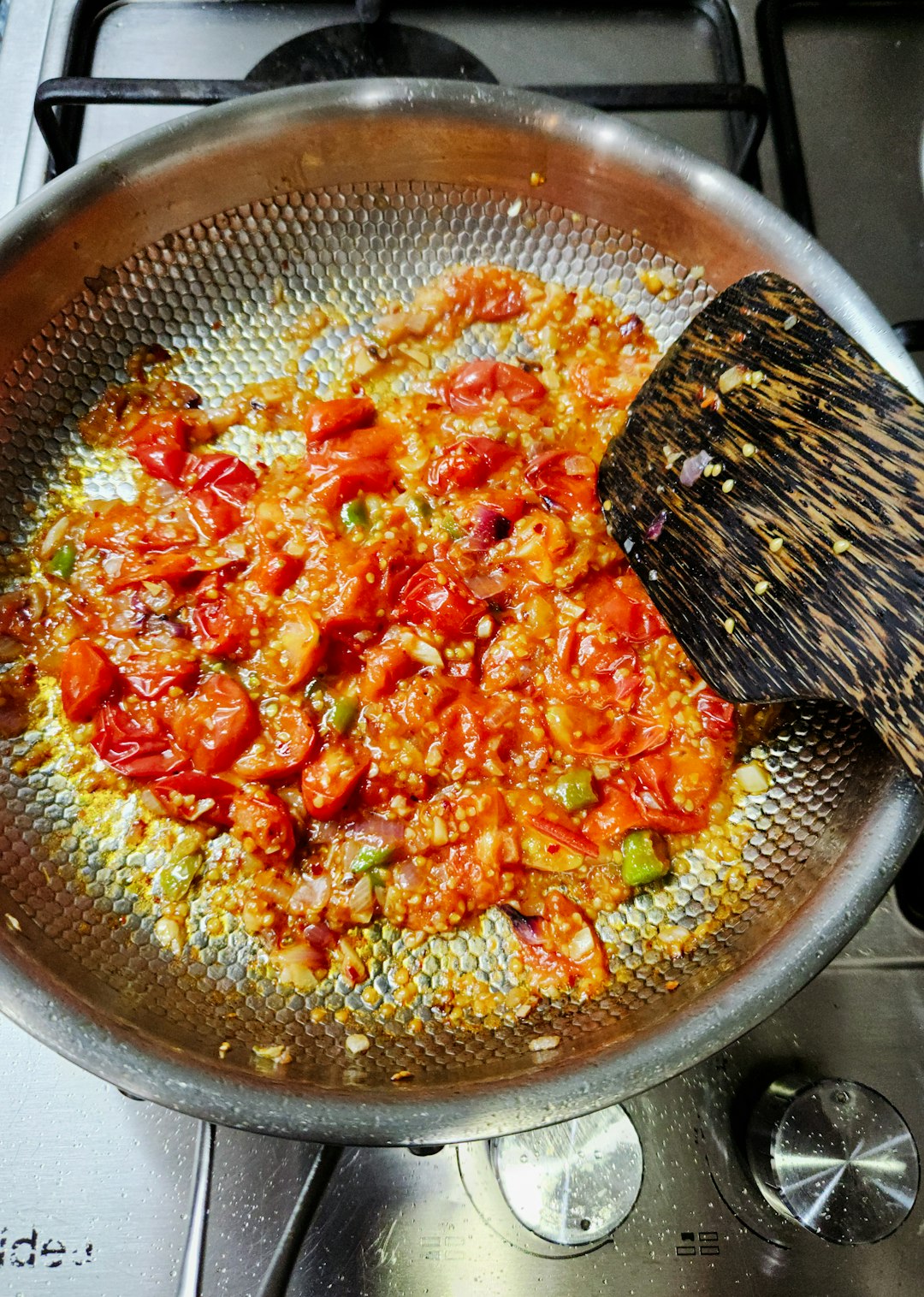 Ingredients being sautéed in a textured stainless steel pan on a stovetop. Chopped tomatoes, onions, and green chilies are simmering in oil, glistening with spices. This mixture likely forms the aromatic base (masala) for a Sri Lankan or South Asian curry or dish.